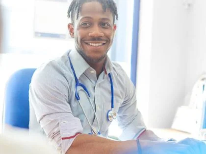 smiling male doctor with stethoscope around neck sat at desk in sunlit room