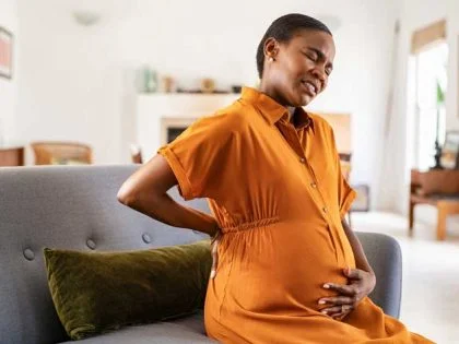 pregnant woman in orange maternity dress rubs her lower back while sitting gret sofa