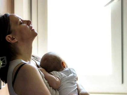 woman with newborn baby held to her chest relaxes by window.