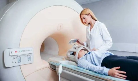 female medic in white coat attending male patient in light blue shirt and dark trousers in front of an MRI Scanner