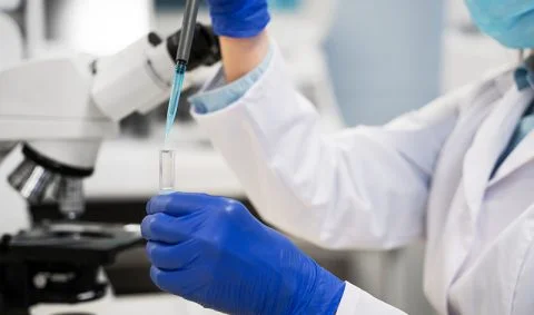 practitioner in white lab coat, blue gloves and wearing mask adds liquid to a test tube infront of a microscope.