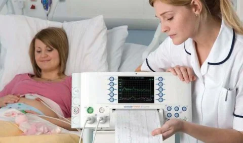 female nurse in white tunic with navy piping examining a monitor printout with woman patient in pink top lying in hospital bed