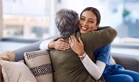 female nurse in blue scrubs hugs elderly patient on couch