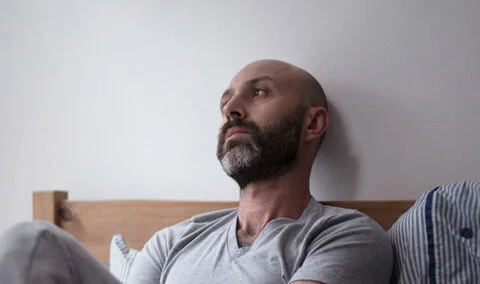 Man with dark beark and grey t shirt sitting on bed and staring into space