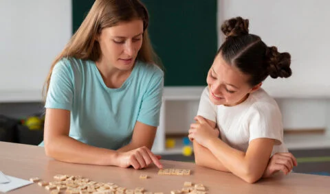 Autism Speech Therapy - therapist using wooden letters to teach girl at tabel in a bright room with large screen TV on wall