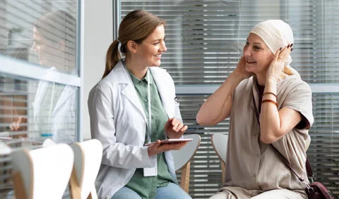 Changing Story of Cancer - female doctor in white coat holding electronic tablet talking to woman patient in windowed room