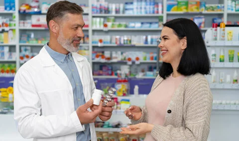 white coated male pharmacist handing pills to a female customer infront of pharmacy shelves