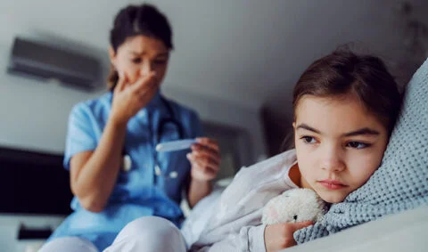 female nurse in blue scrub tunic standing behing young girl lieing on a bed with cuddly toy.