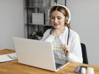 woman sitting at desk with laptop holding packet of yellow pills and rubbing her throat